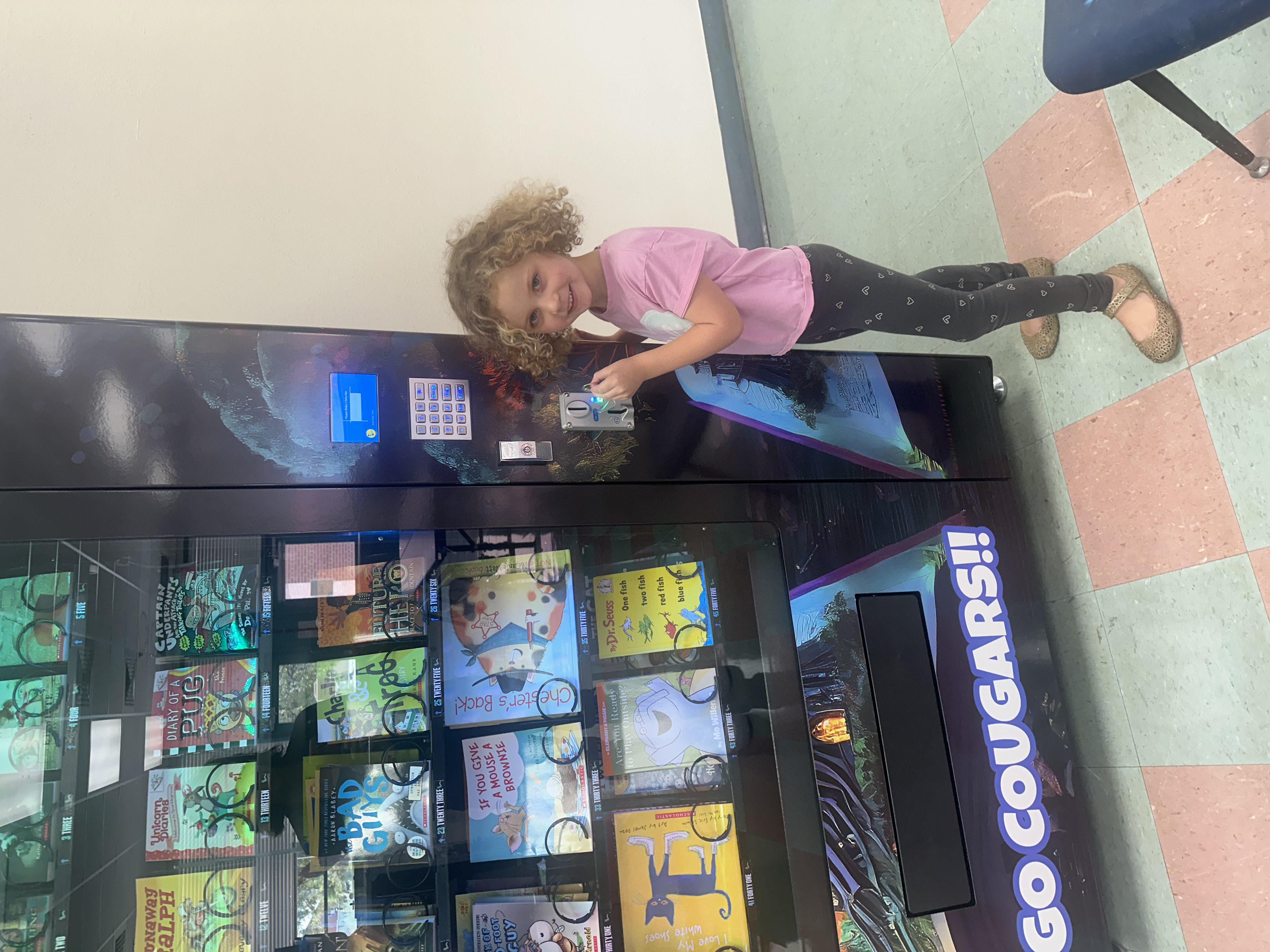 A little girl with curly blonde hair puts her token into the book vending machine