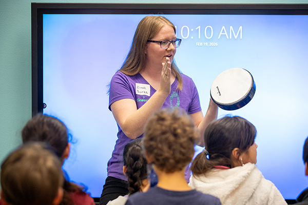 A woman holds up a drum and plays for students