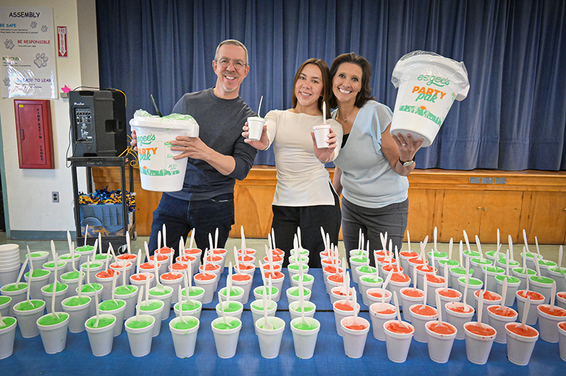 A man and woman, left and right, hold giant containers of eegees while a woman, center, holds two cups behind a table filled with green and red eegees
